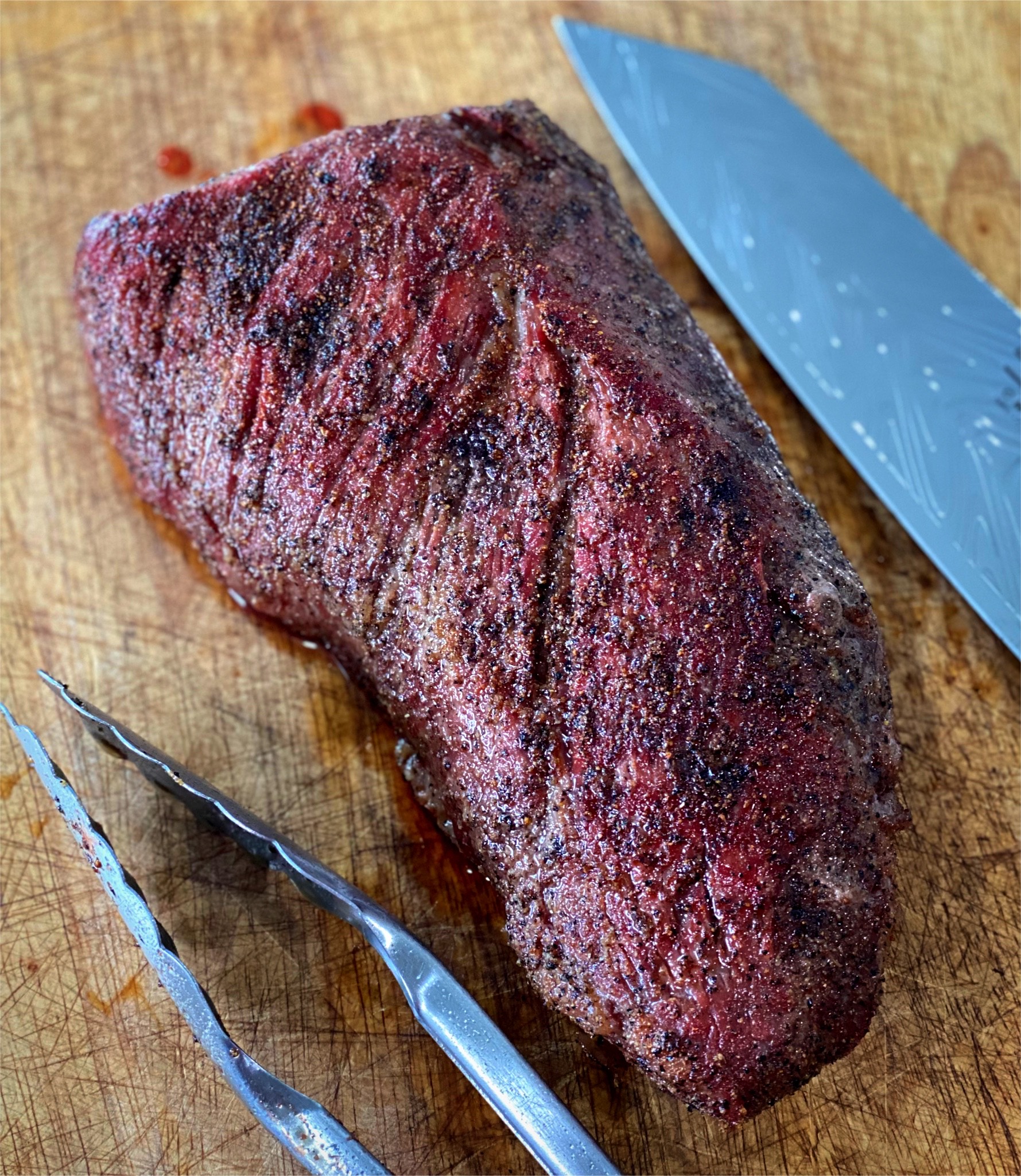Smoked tri tip resting on the cutting board by a knife and tongs.